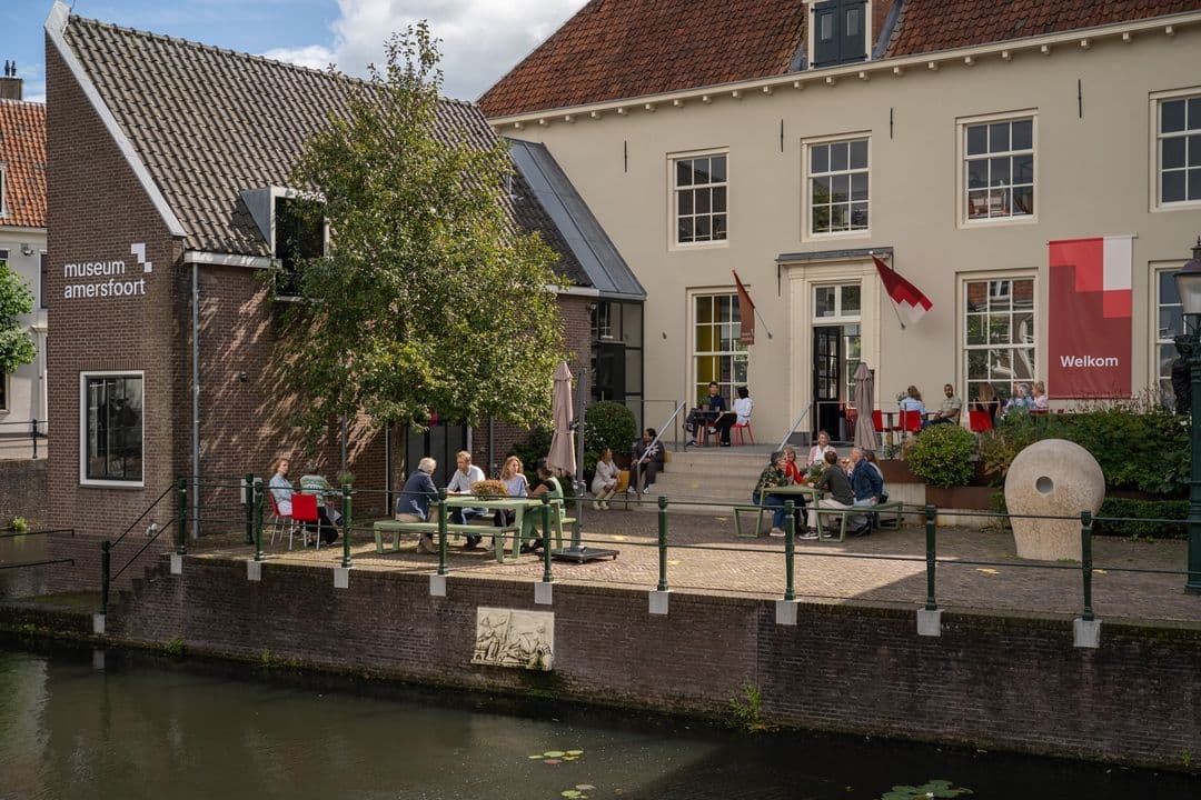 Foto van het gebouw. Op het terras, aan de kade zitten bezoekers. Ze praten, drinken een drankje en geniet samen.